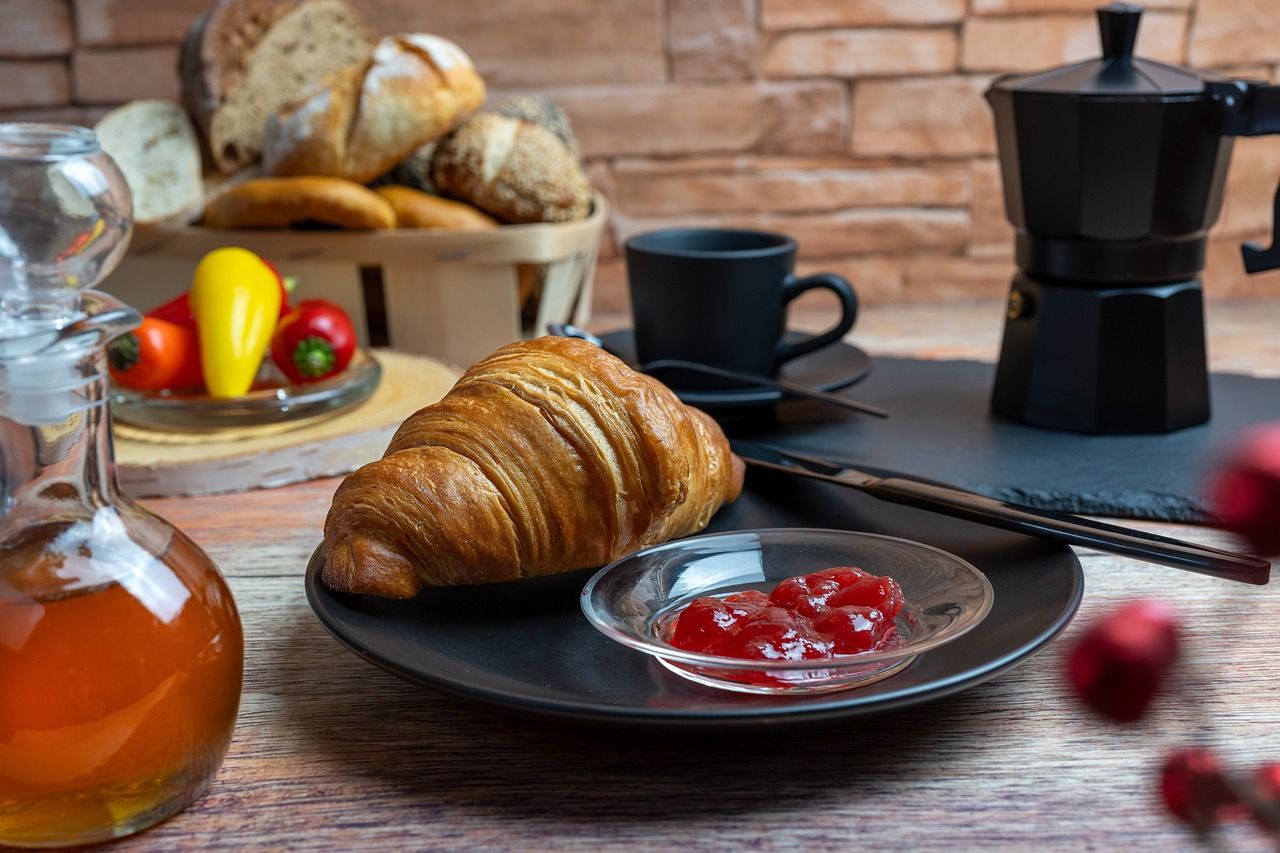 Mesa de café da manhã com croissant, geleia, cesta de pães e cafeteira italiana