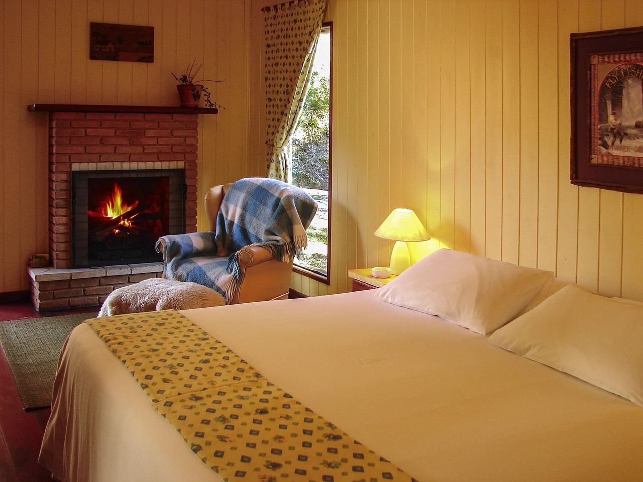 Quarto do chalé com lareira de tijolos acesa, poltrona com manta xadrez e cama de casal