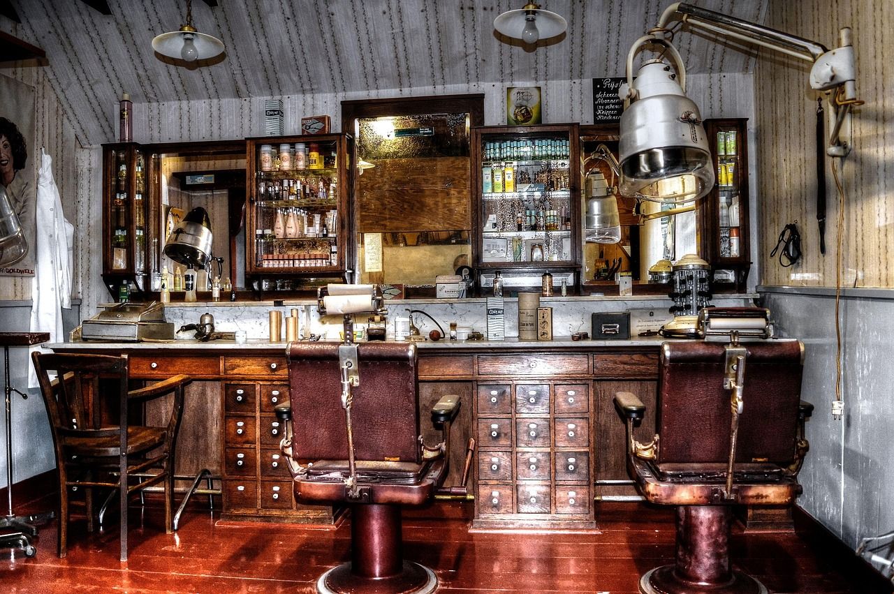 Salão de barbearia antiga, com duas cadeiras de couro, bancada de madeira e espelhos