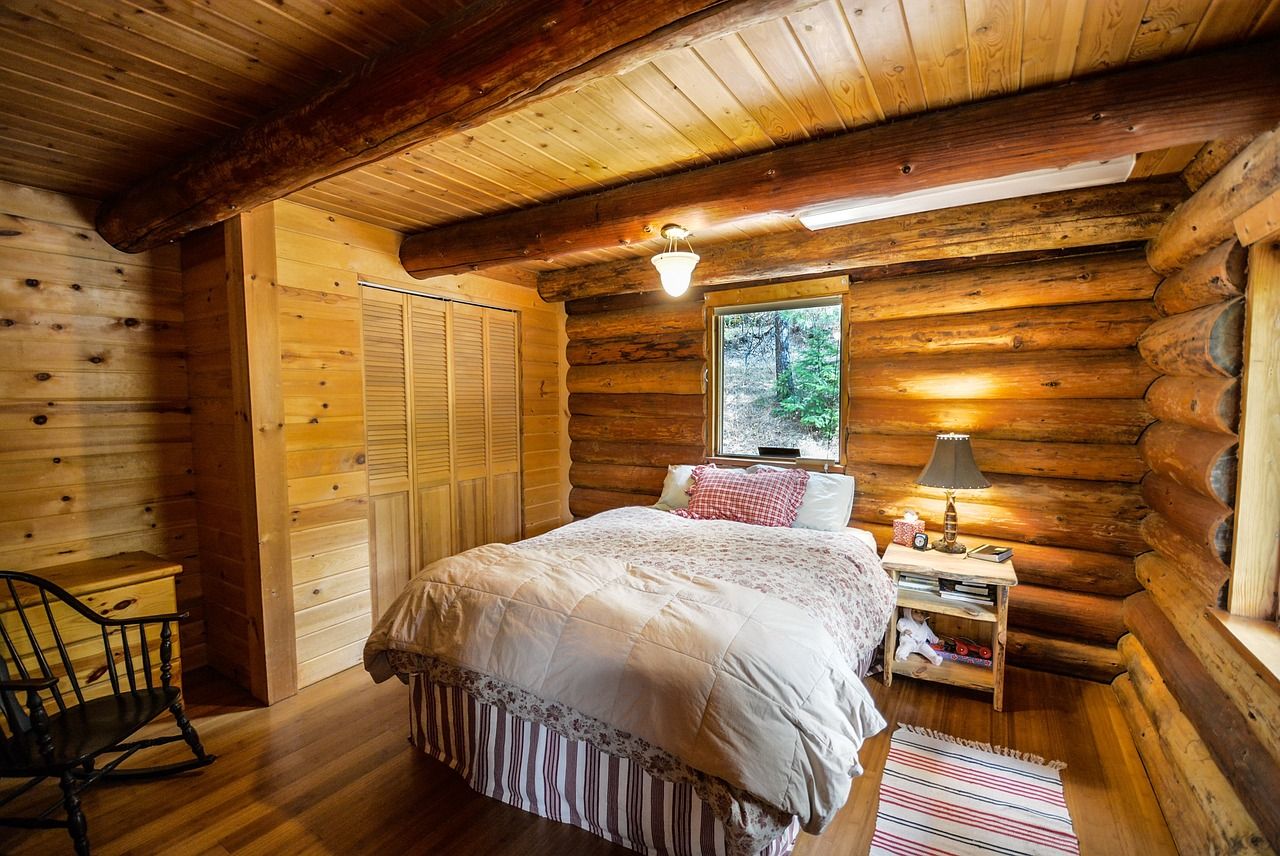 Quarto com paredes de tora, cama de casal com colcha clara, abajur aceso e janela para o bosque