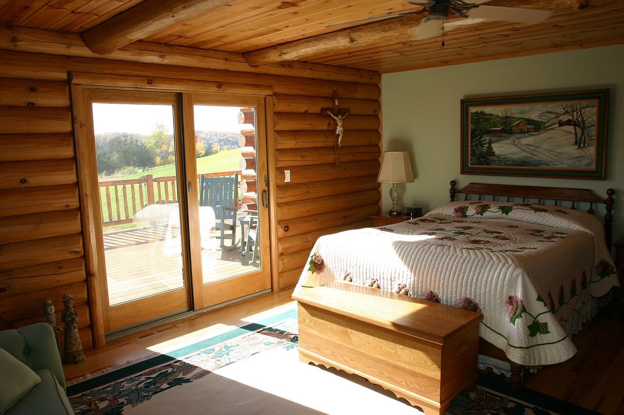 Quarto de madeira com cama de casal, baú aos pés da cama e porta de vidro aberta para a varanda